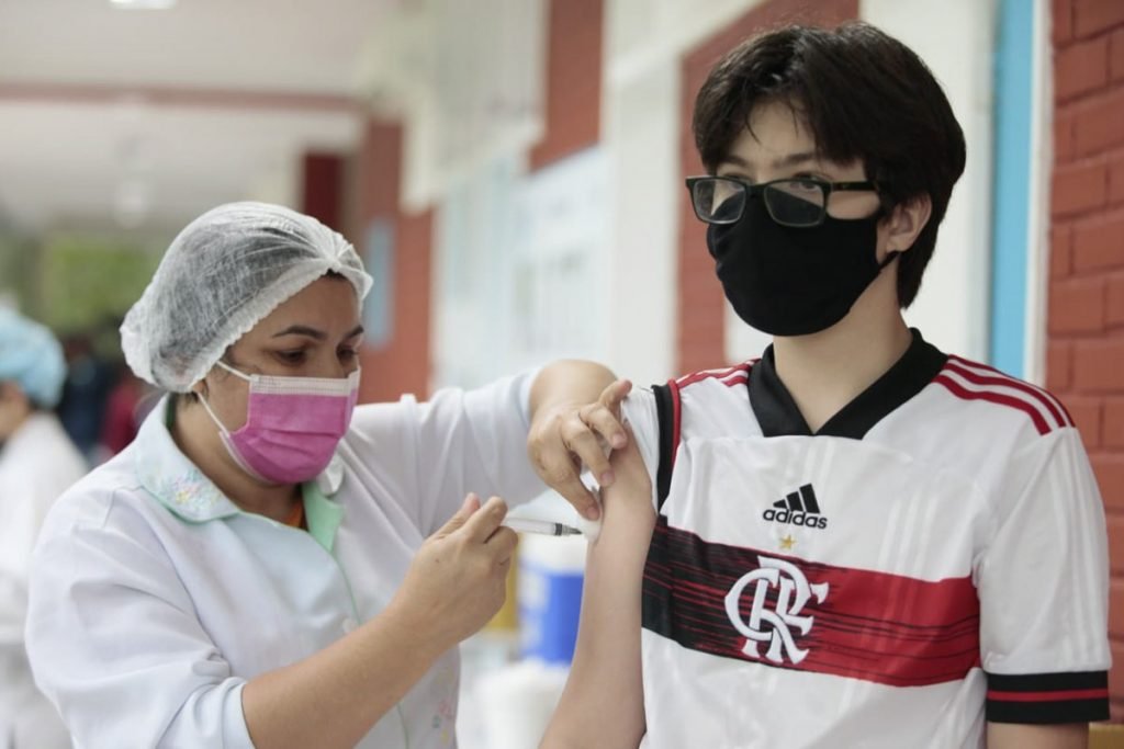 Adolescentes são vacinados no Rio de Janeiro com segunda dose. Na foto, jovem branco, com camisa do Flamengo, óculos de grau e máscara preta recebe dose de uma enfermeira branca, de jaleco e touca branca e máscara rosa