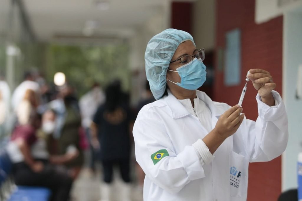 Vacinação na cidade do Rio de Janeiro segue intensa para imunizar toda a população contra o Covid-19.  Na foto, uma enfermeira branca, com jaleco e touca, óculos de grau e máscara branca segurando uma seringa de vacina