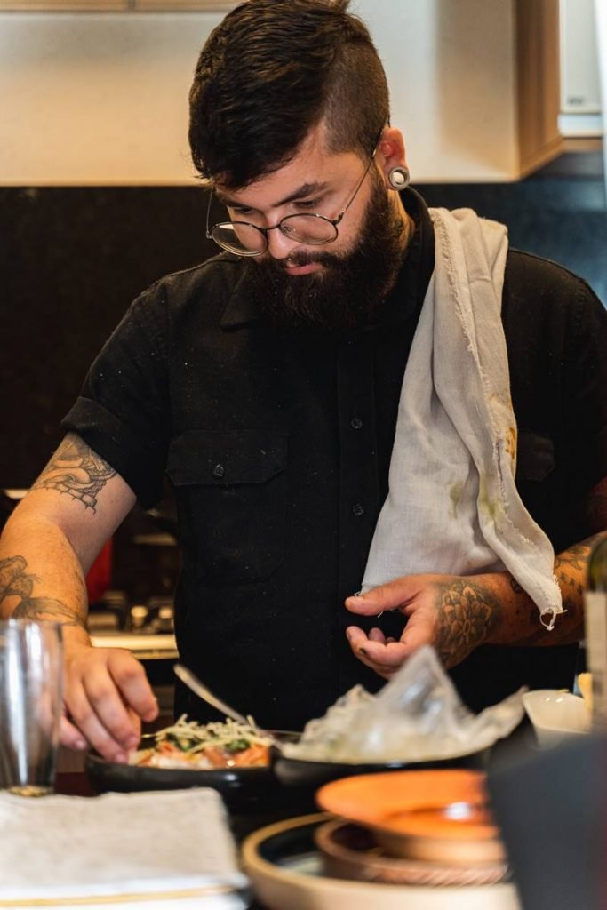 Na foto, um homem barbudo e branco com roupa preta está arrumando um prato de comida