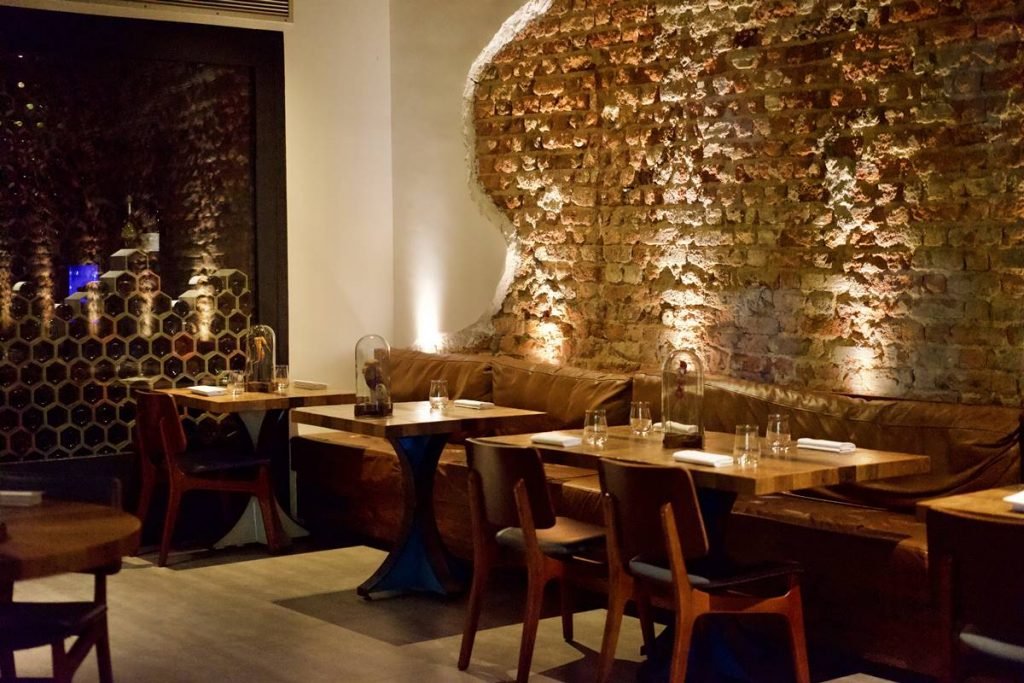 Na fotografia vemos uma sala de jantar em um restaurante. A parede em textura de tijolinhos, sofás e mesas e cadeiras de madeira