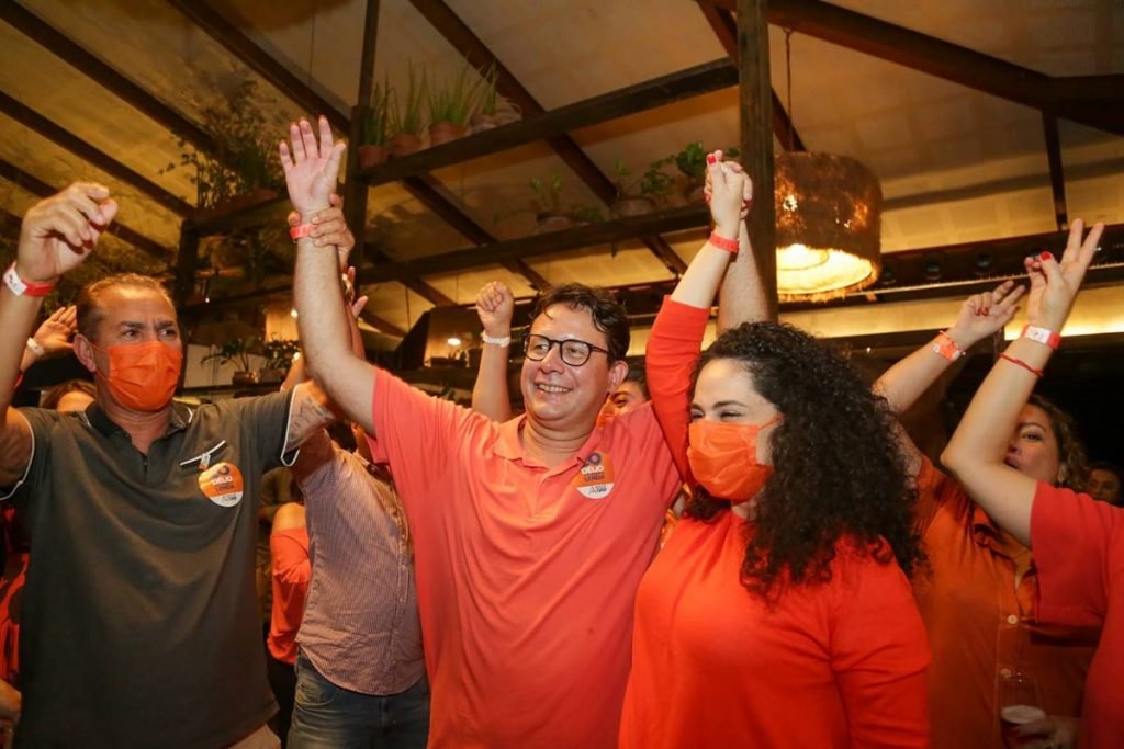 Homem de laranja com mãos levantadas em comemoração e uma mulher ao lado também de laranja de cabelo castanho cacheado