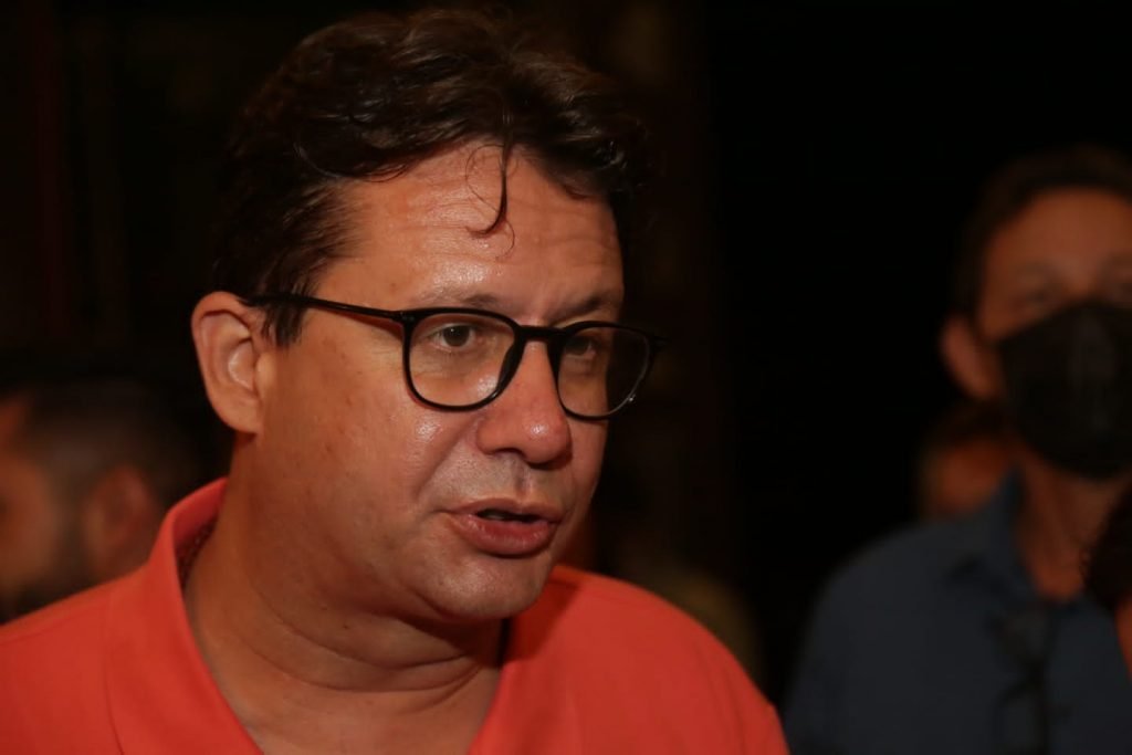 homem branco de camisa laranja dando entrevista