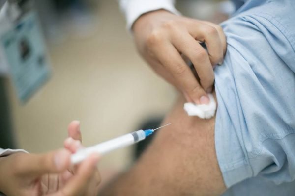 Na imagem colorida uma mão aplica seringa com vacina em braço