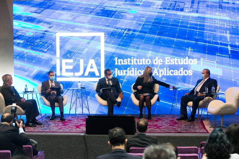 Pessoas sentadas em cadeiras sobre um palco e com telão