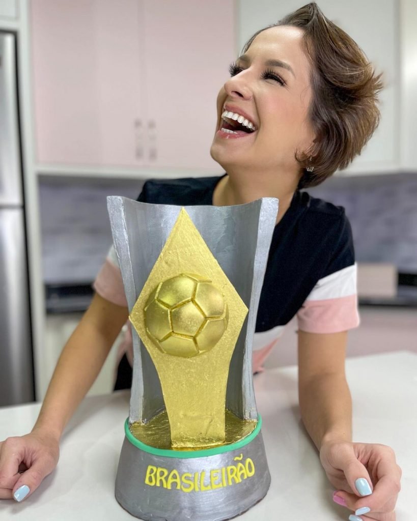 Na imagem, uma mulher sorridente posa em frente ao troféu do brasileirão feito de bolo