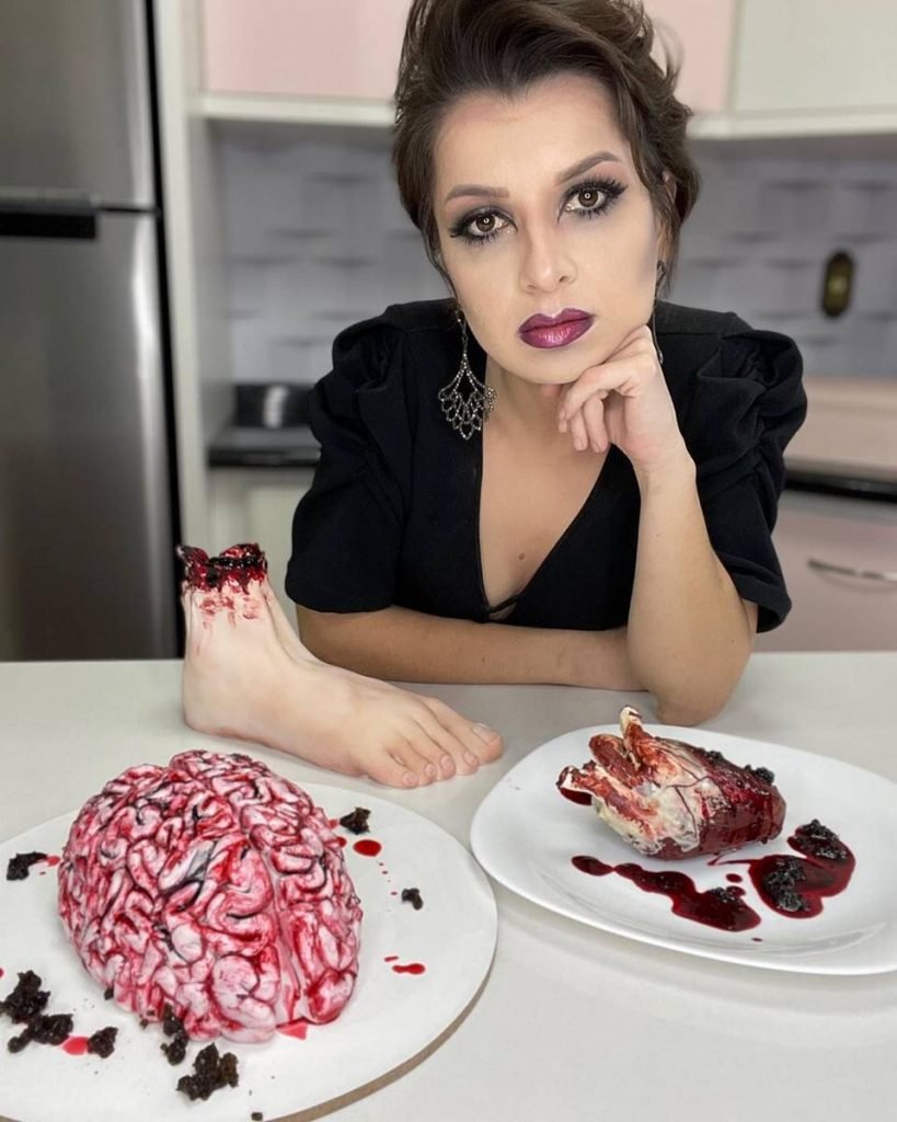 Na imagem, uma mulher maquiada para Halloween posa em frente a bolos em formato de partes do corpo humano com sangue