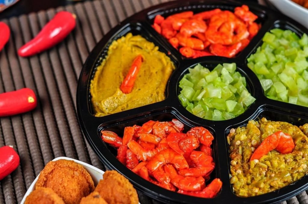 Na foto, um recipiente com diversas divisórias contendo itens de acarajé