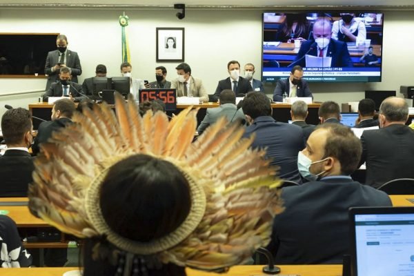 Homem indígena em sala do Congresso, enquanto, ao fundo, de terno, deputados e ministros conversam