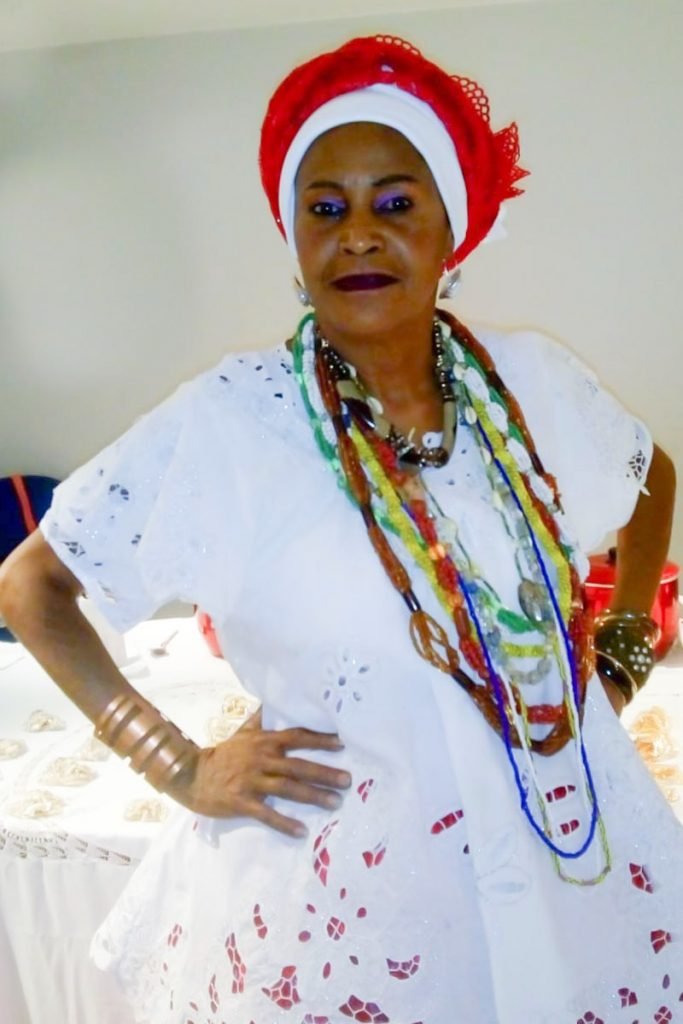 Na foto, uma mulher negra com vestes brancas de baiana e colares coloridos