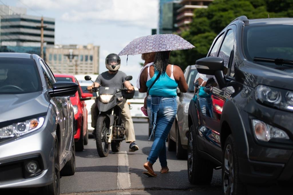 Mulher com blusa azul e guarda0chuva lilás em meio a carros. Ao fundo um homem utilizando capacete em cima de uma motocicleta