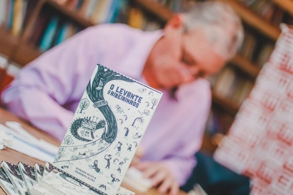Na foto vemos em primeiro plano a capa de um livro. Ela é branca com desenhos verdes. Ao fundo, desfocado, um homem de camisa roxa escreve