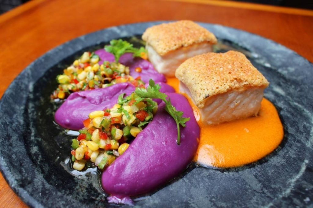 Na foto, prato cinza com pescada em crosta de castanhas brasileiras, creme de moqueca, mousseline de batata doce roxa e vinagrete de milho tostado