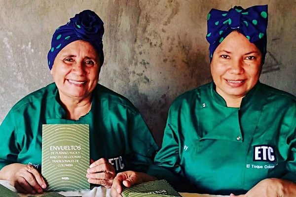Na foto temos duas mulheres vestidas com dolmãs verdes e turbantes azuis escuros. A da esquerda é uma senhora e segura um livro d capa verde e a outra aparenta uns 40 anos. As duas sorriem