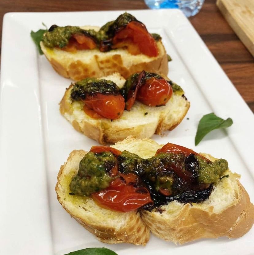 Na foto, prato branco om Trio de bruschetta de tomates assados com pesto de manjericão, castanha de caju e redução de balsâmico