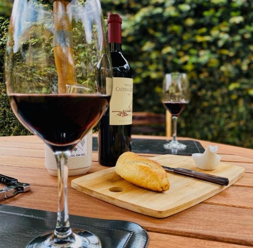 Na imagem, uma taça de vinho pela metade, ao fundo um queijo cortado em uma tábua de madeira e uma garrafa de vinho