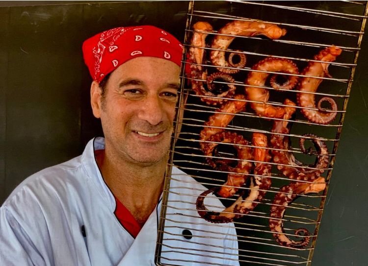 Na foto, um homem com bandana vermelha e roupa branca segura uma grelha com tentáculos de polvo grelhados