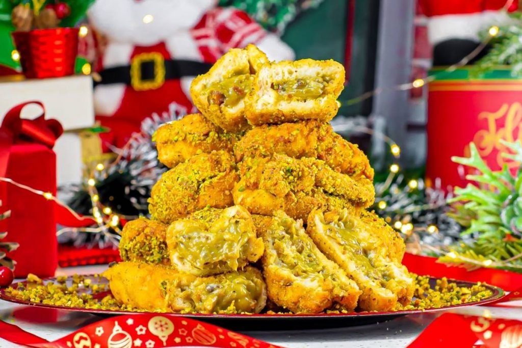 Na foto temos uma bandeja com varias rabanadas recheadas com creme verde empilhadas e decoração de natal ao fundo