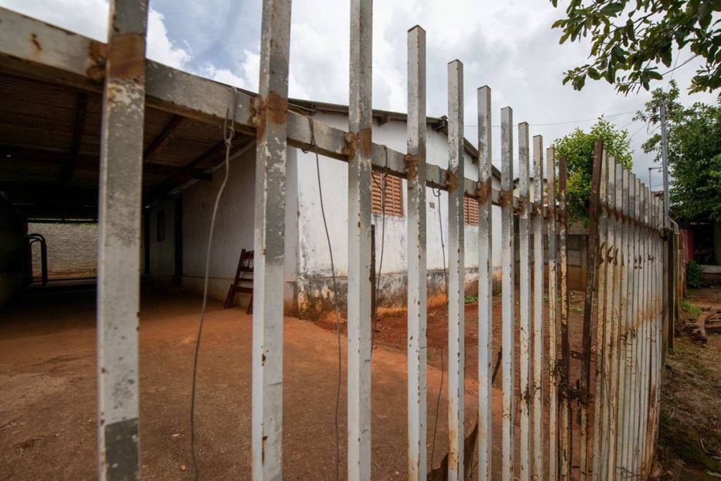 Portão branco, com partes enferrujadas, e uma casa branca com duas janelas marrons atrás