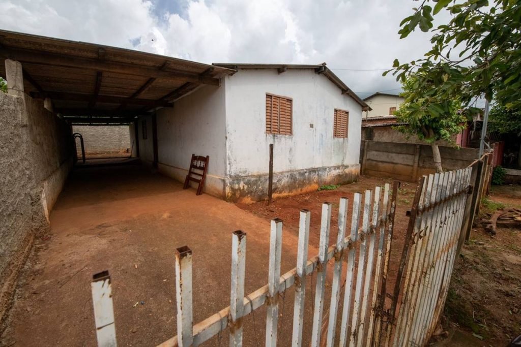 Casa com parede branca e duas janelas marrom. Na frente, um portão branco, mas enferrujado.