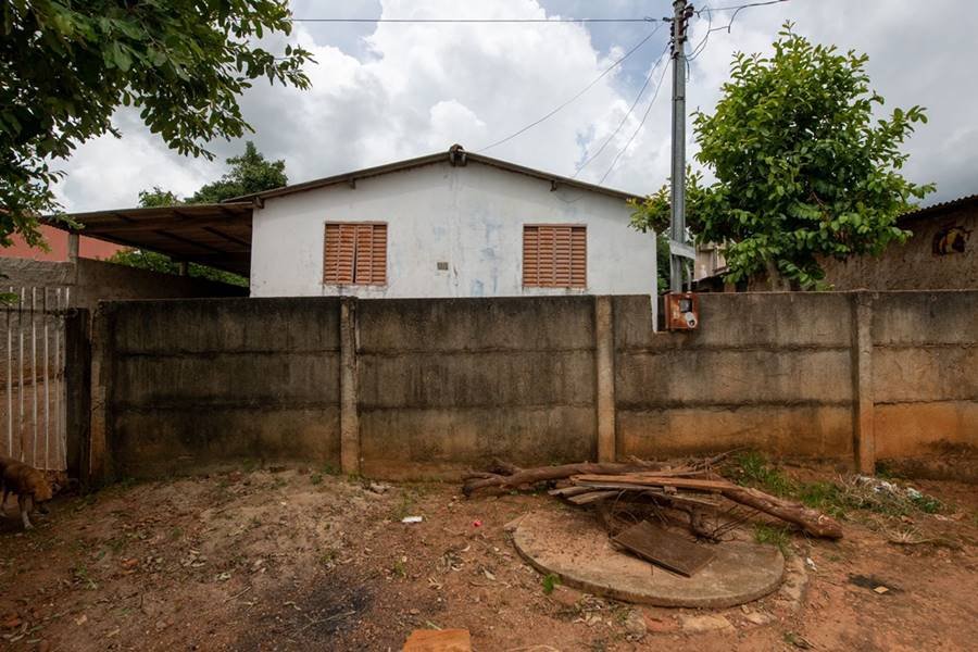 Há três planos na imagem. A primeira mostra um terreno baldio com terra e sujeiras. O segundo é um muro sujo de barro. O terceiro é uma casa de parede branca e com duas janelas em laterais opostas.