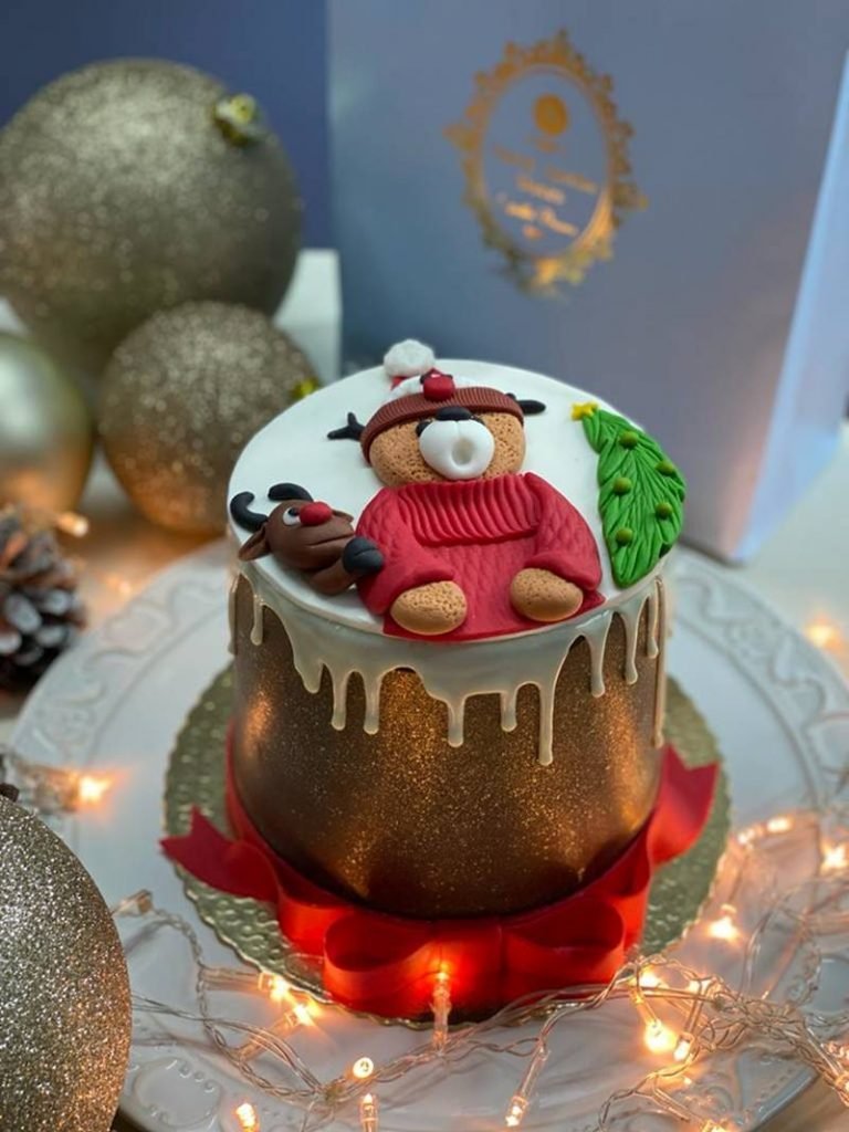 Na foto temos um chocotone coberto com chocolate e brilhos. Por cima cai uma calda branca e existe um urso de blusa de frio vermelha feito de massinha, uma rena e uma folha com bolinhas colorda