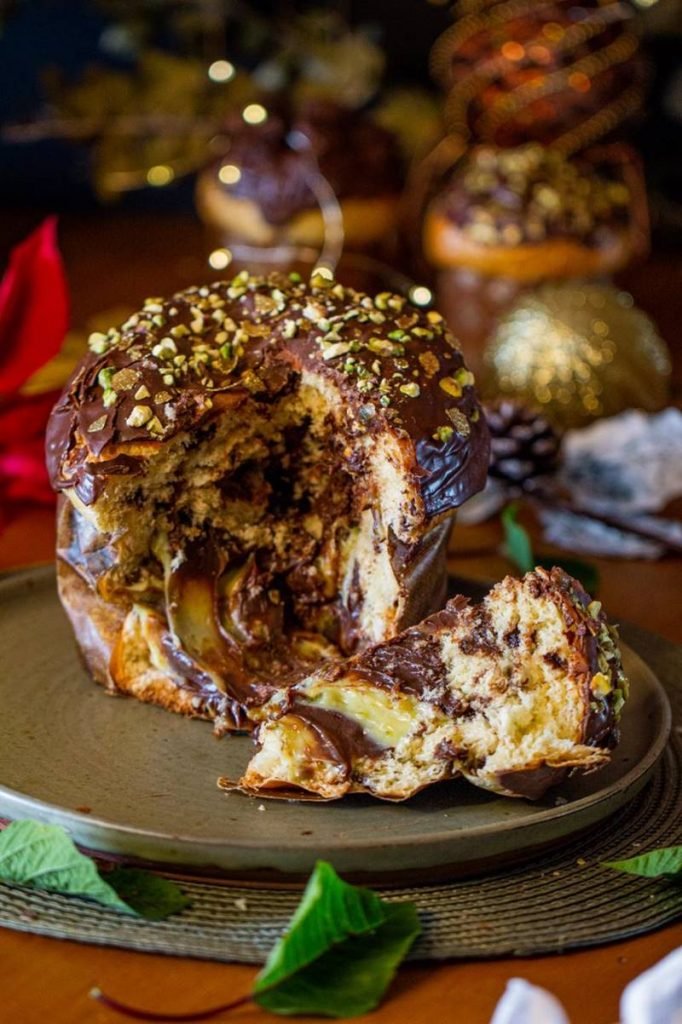 Na foto vemos um chocolate recheado faltando uma fatia. A fatia esta ao lado dele sobre um prato redondo verde. O recheio é marrom e verde. Ao fundo, decorações de Natal
