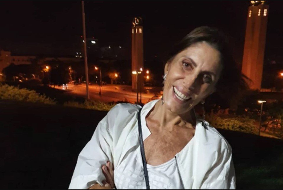 Na fotografia colorida, Mila Moreira aparece à esquerda. Ela veste roupas brancas e sorri para a câmera