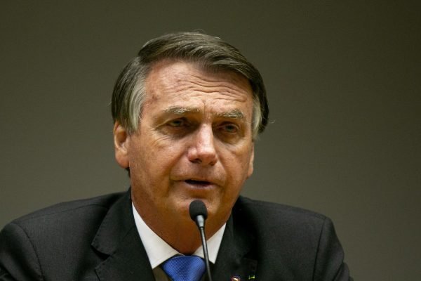 Fotografia colorida. Bolsonaro aparece no centro da imagem falando ao microfone. Ele veste um paletó com gravata azul e aparenta estar preocupado