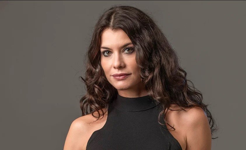 Na imagem colorida, a atriz Aline Moraes aparece centralizada. Ela usa blusa preta com ombros à mostra. Os cabelos ondulados de Aline possuem tom achocolatado e caem sobre os ombros dela. Ela olha séria para a câmera.
