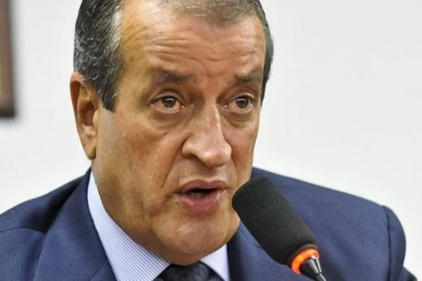 Fotografia colorida. Valdemar aparece no centro da imagem falando ao microfone. Ele veste paletó azul e gravata da mesma cor