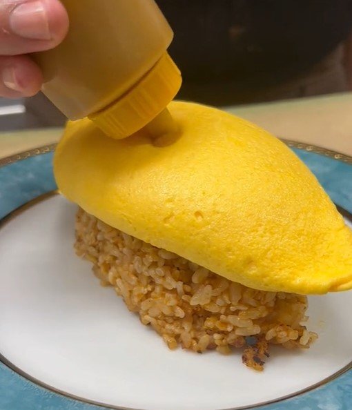 Na foto, uma pessoa injeta um líguido amarelo dentro de um omelete fechado que está em cima de um arroz marrom