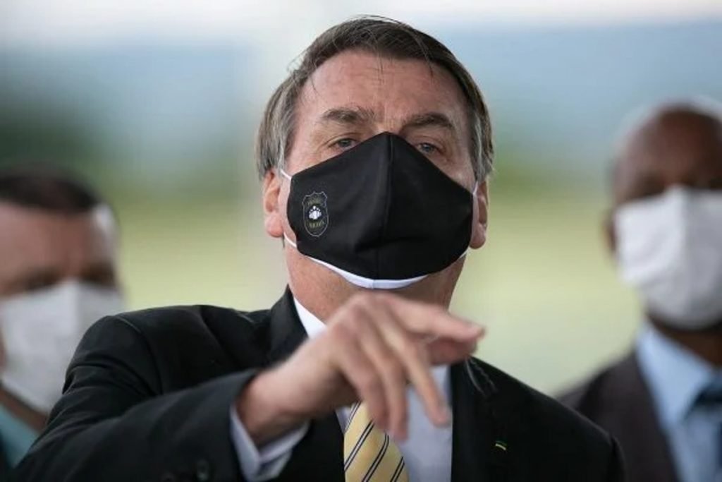 Fotografia colorida. Jair Bolsonaro aparece no centro da imagem usando máscara preta e apontando para alguém em sua frente