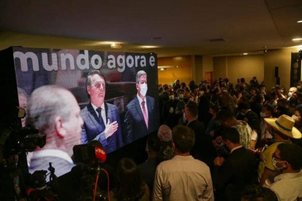 Fotografia colorida.  Bolsonaro aparece no telão do local, mão um paletó, com a peito em sina de respeito ao Hino Nacional