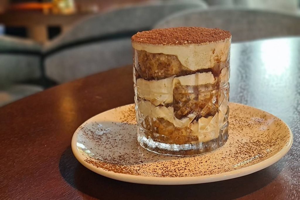 Na foto vemos um prato branco com cacau salpicado e um copo de vidro com um doce em camadas de biscoito e creme branco. Ao fundo, uma poltrona cinza