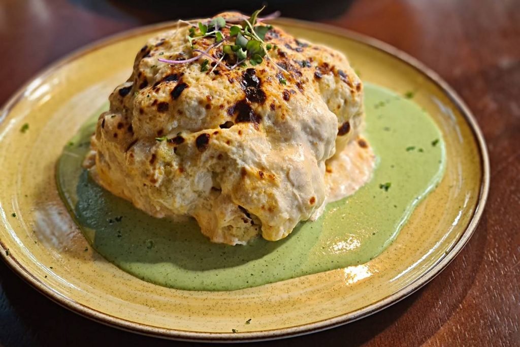 Na foto vemos um branco amarelado com um creme verde e o centro um preparo quadrado gratinado, com tons amarelos e pretos e folhas verdes por cima