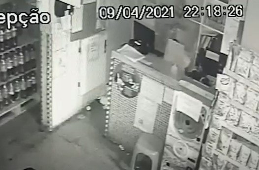 Em uma imagem de câmeras, preta e branca, um homem mexe ao que parece, atrás de um balcão, em uma caixa registradora