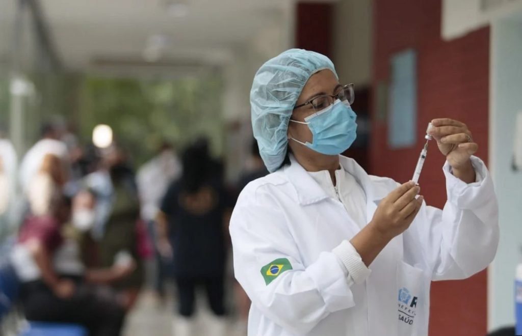 Na imagem colorida, uma profissional de saúde está posicionada ao lado direito. Ela usa jaleco branco, touca e máscara na cor azul e segura uma seringa nas mãos