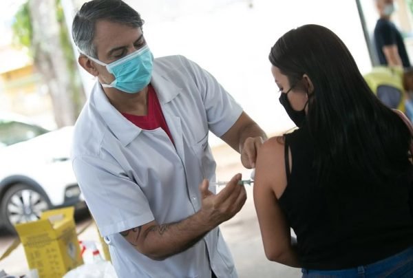 In the color image, a health worker is positioned on the left and a woman on the right.  He wears a white coat, a blue mask and holds a syringe in his hands.  She wears a black blouse, wears a dark mask, and has long, black hair.