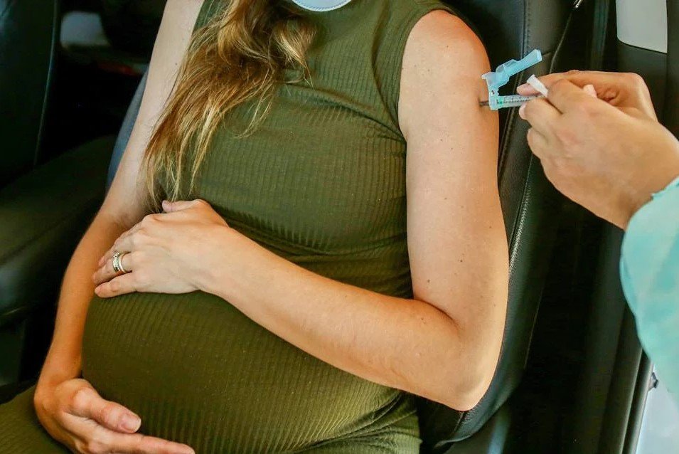 Na imagem colorida, uma grávida aparece posicionada no centro. Ela usa vestido verde e está com as mãos em cima da barriga. Na parte direita da imagem, há duas mãos segurando uma seringa