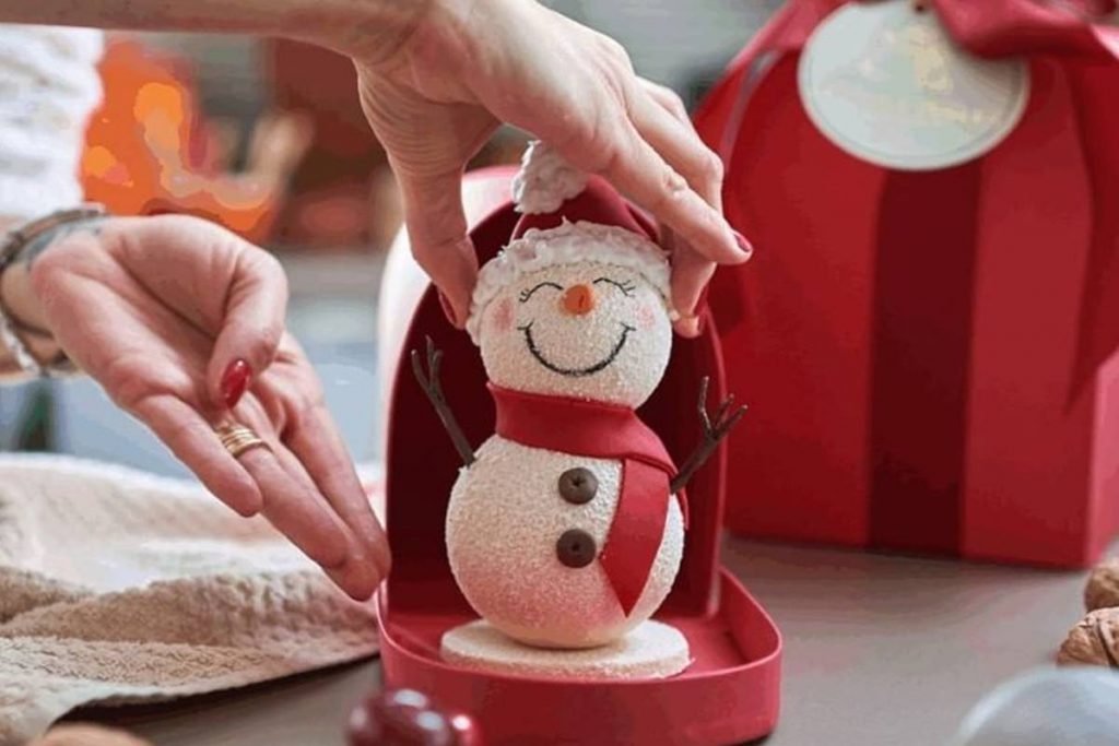 Na foto temos duas mãos segurando um boneco de neve que usa um cachecol vermelho e um gorro vermelho