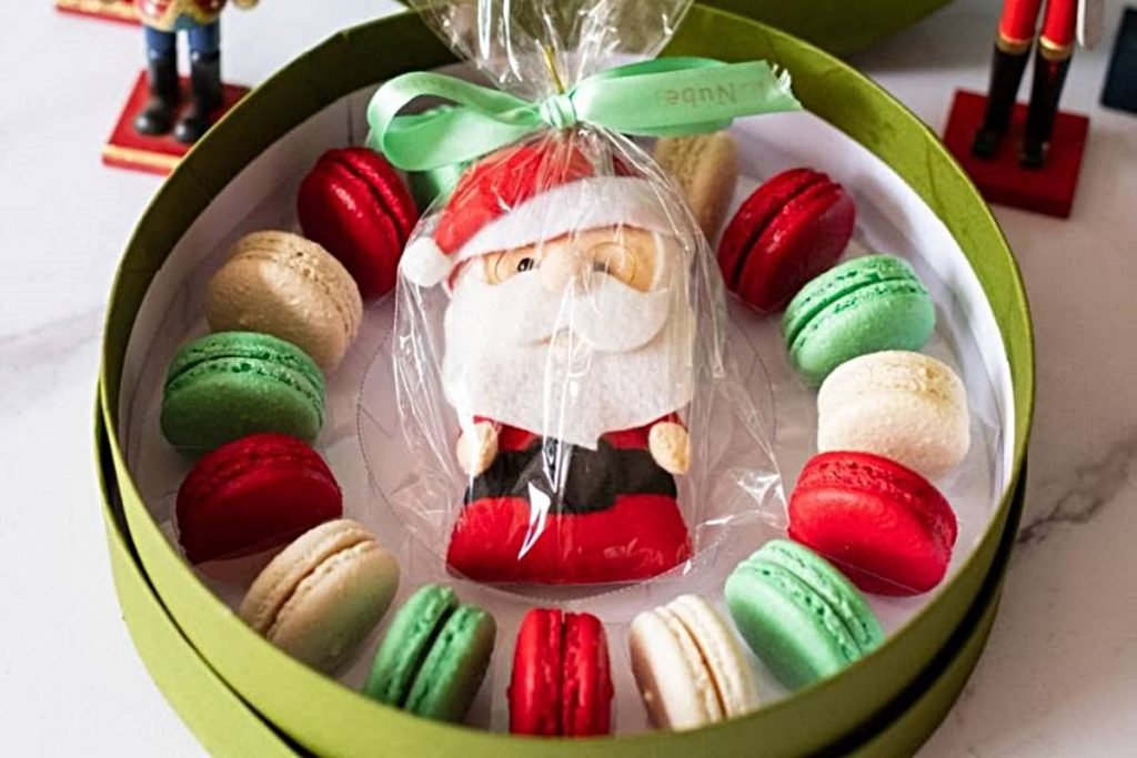 Na foto temos uma caixa verde por fora e branca por dentro com macarons vermelhos, branos e verdes dispostos em formato circular e com um papai noel de feltro no meio