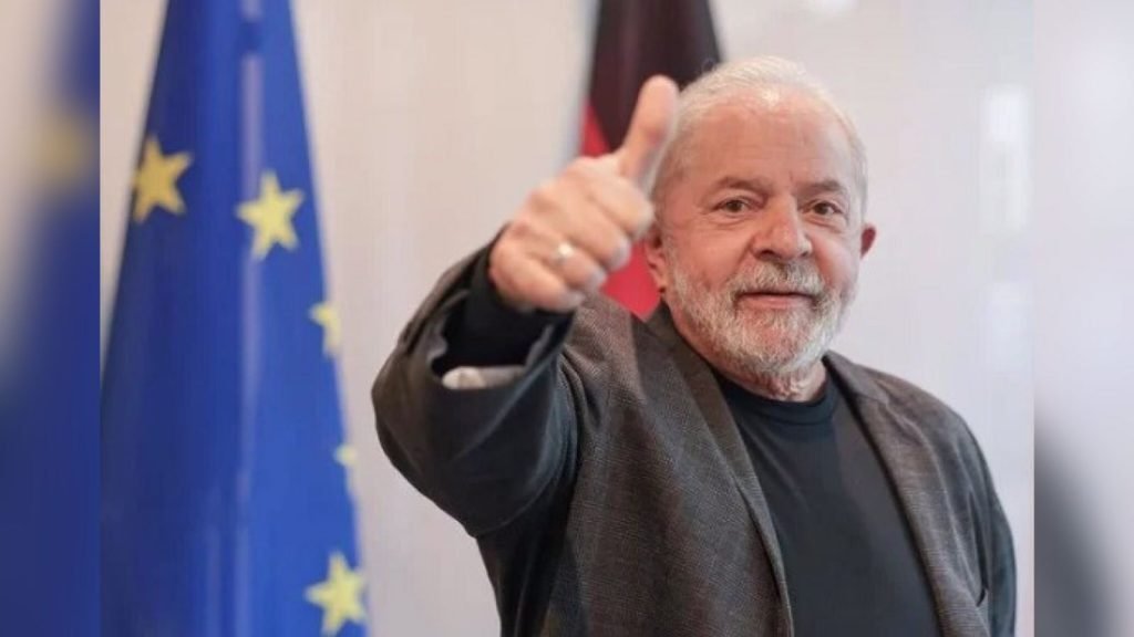 Na imagem, Lula está posicionado do lado direito da imagem.  Ele usa blazer e camiseta escura, posicionando a mão fazendo sinal de 