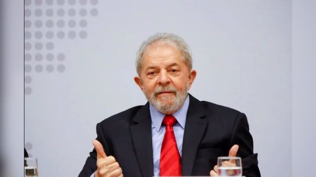Na imagem, Lula aparece centralizado.  Ele usa blazer escuro, camiseta azul e gravata vermelha.  Há um copo ao lado dele e suas mãos estão para cima e fazendo o sinal de 