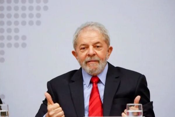 Na imagem colorida, Lula aparece centralizado. Ele usa blazer escuro, camiseta azul e gravata vermelha. Há um copo ao lado dele e suas mãos estão para cima e estão fazendo o sinal de jóia