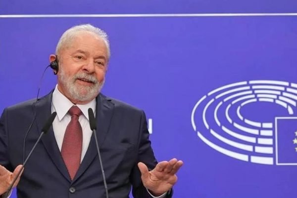 Na imagem colorida, Lula está posicionado do lado esquerdo, sob fundo azul. Ele usa terno escuro, camiseta branca e gravata vermelha. Na frente dele há um microfone e ele está com as mãos levantadas - Metrópoles