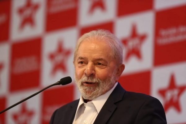 Na imagem colorida, Lula encontra-se centralizado. Ele usa blazer preto e camisa branca. Na frente dele há um microfone e na parte de trás a um fundo vermelho e com a imagem do PT