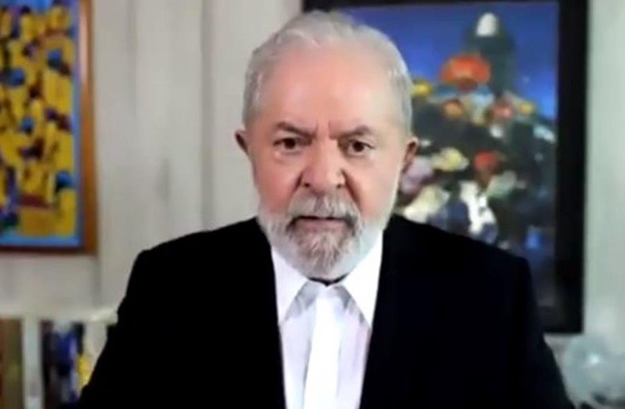 Na imagem colorida, Lula está posicionado no centro da imagem. Ele usa terno preto, camiseta branca e olha sério para a câmera