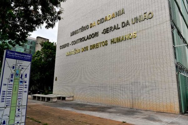 Na imagem colorida, a parte lateral do Ministério da cidadania exibe o letreiro do órgão e está posicionado à direita. Na parte esquerda há folhas de árvores