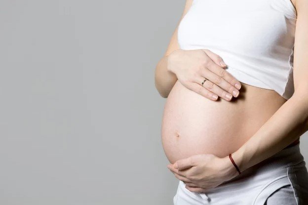 Na imagem colorida, uma gravida está posicionada do lado direito. Ela está com a barriga a mostra e usa blusa e calça branca
