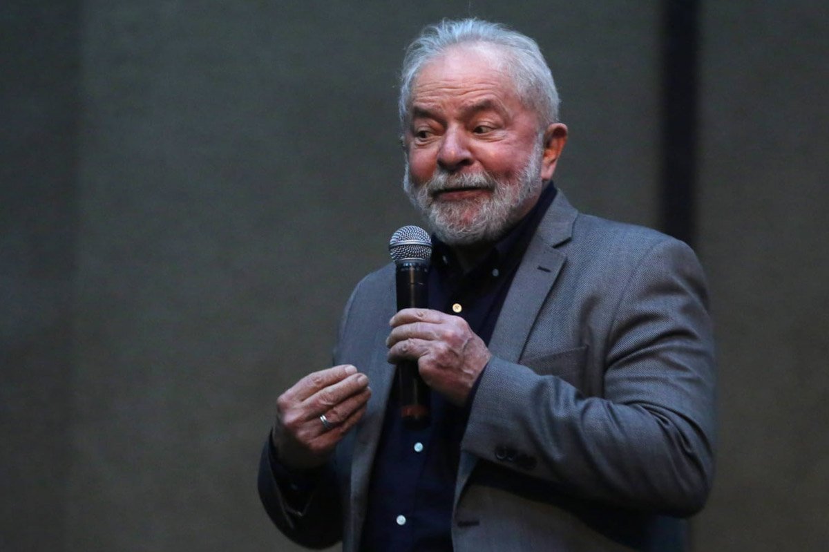 Ex-presidente Lula fala ao público. Ele usa terno e não está com máscara. Segura um microfone e gesticula sob um fundo cinza - Metrópoles